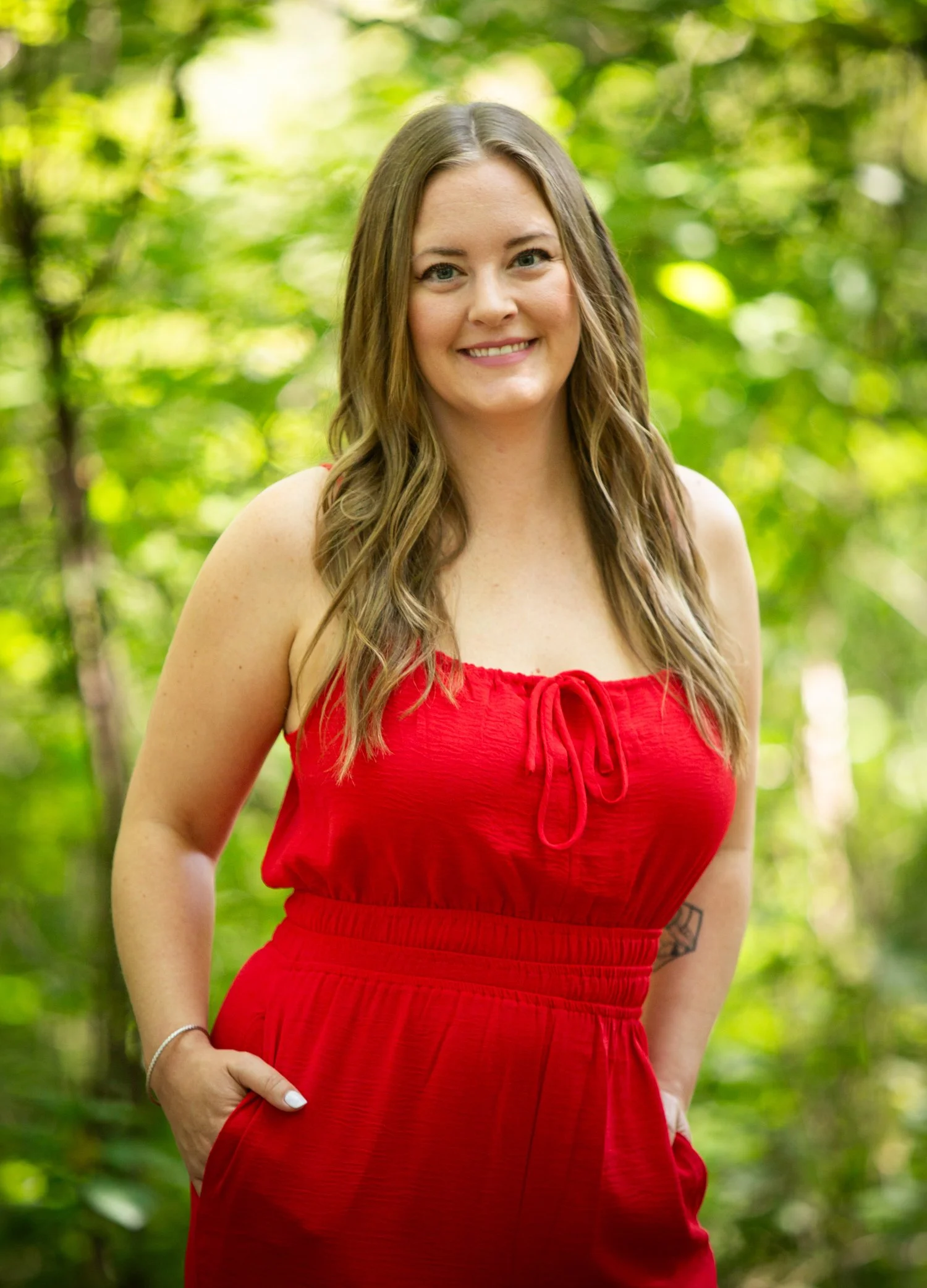 A young woman with long wavy brown hair, smiling, wearing a red sleeveless dress, standing outdoors in a lush green forest.