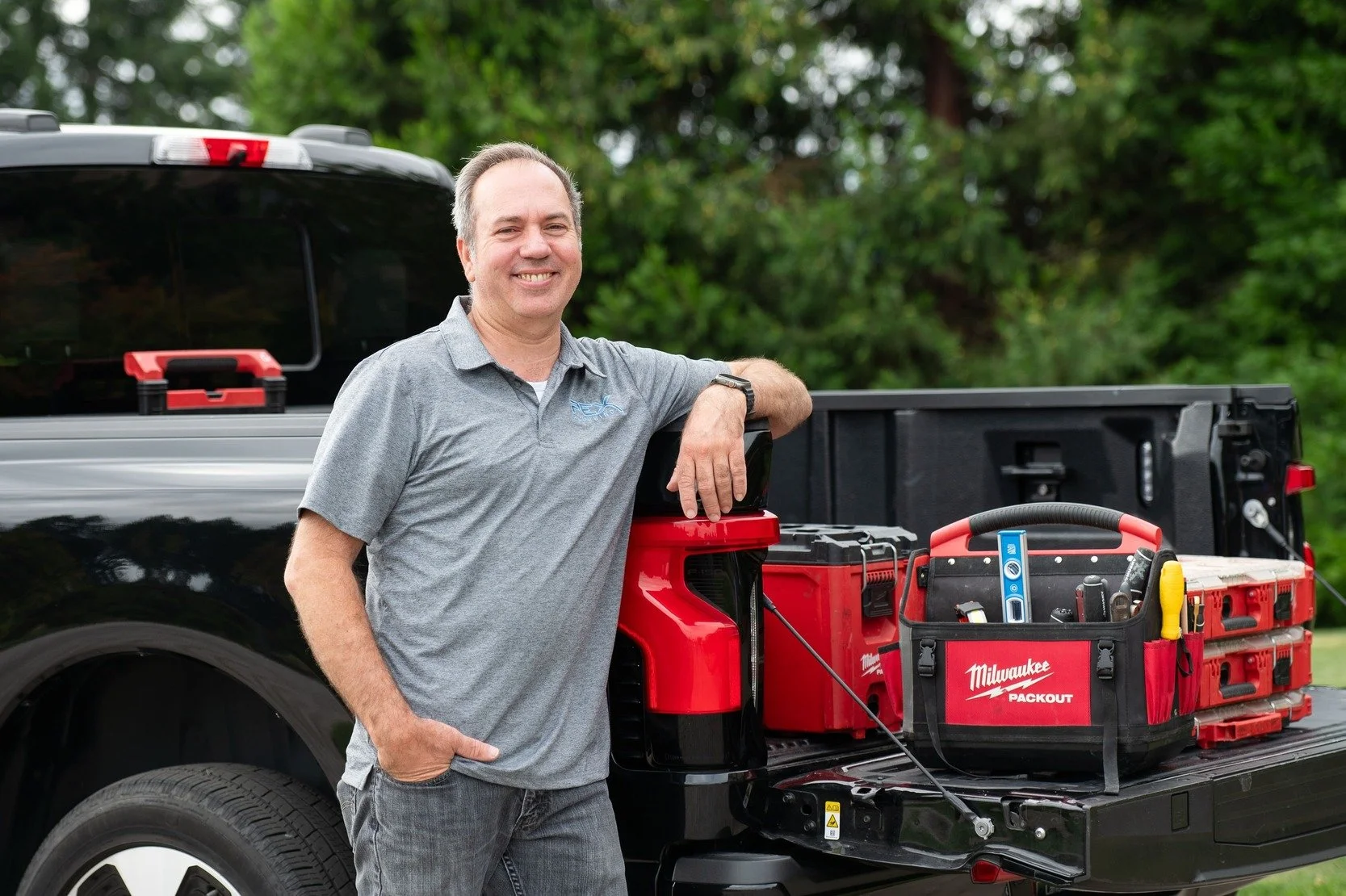 A man in a gray polo shirt leaning on the side of a pickup truck loaded with tools, smiling, with a green and wooded background.