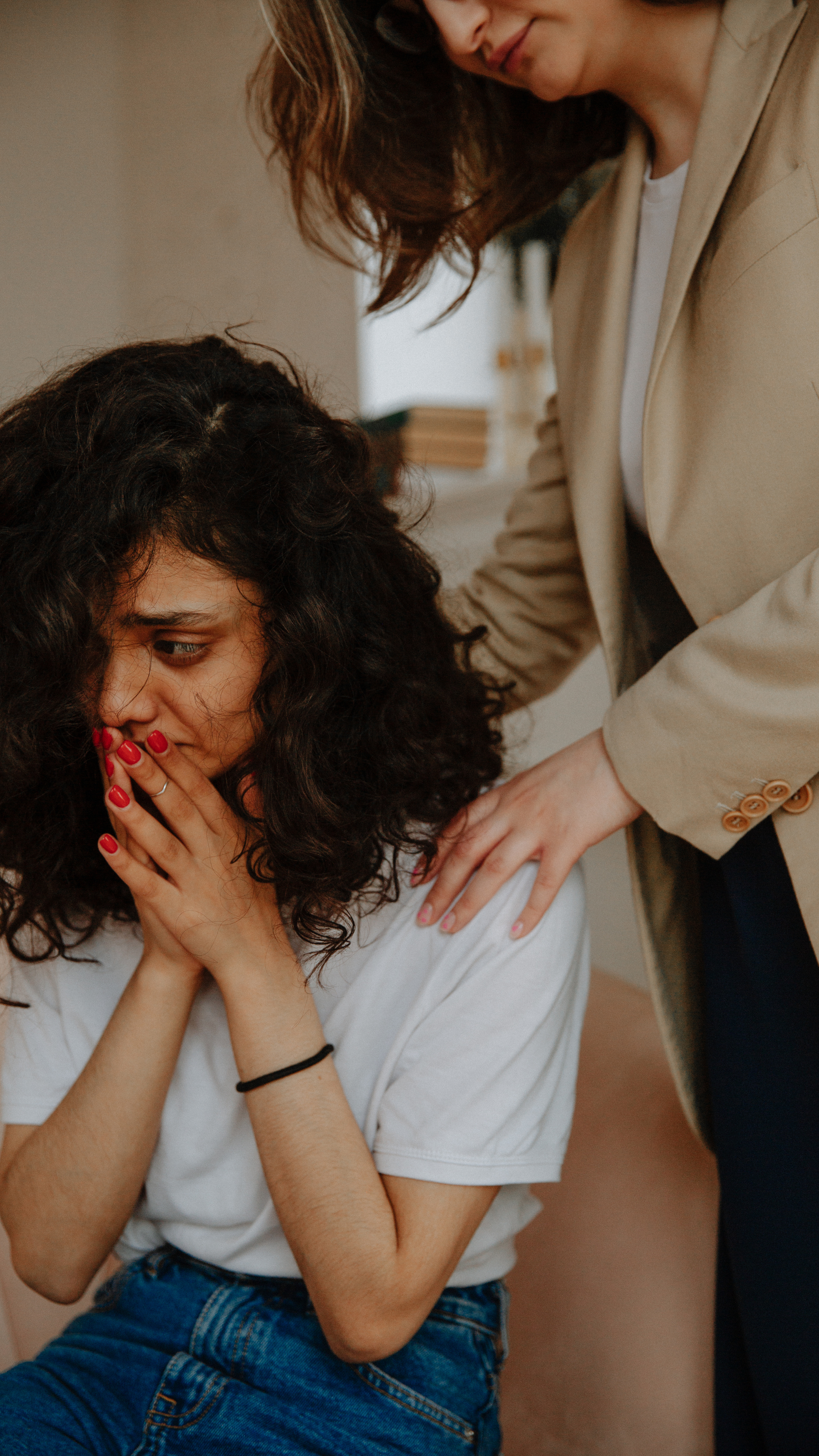 A young woman with curly hair sits with her hands covering her face, her nails painted bright red. She is wearing a white t-shirt and blue jeans. An older woman, possibly a caregiver or nurse, leans over her, gently placing a hand on her shoulder. The setting appears to be indoors, possibly a therapy or counseling session.