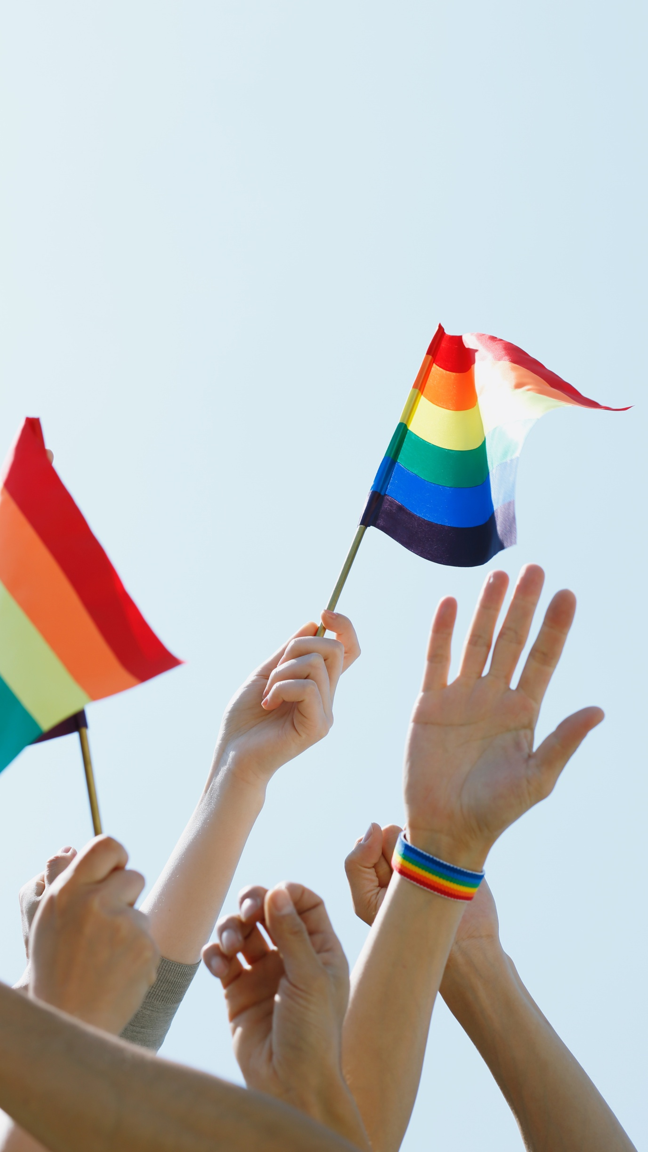 Multiple hands holding rainbow pride flags and raising them in the air against a clear sky.