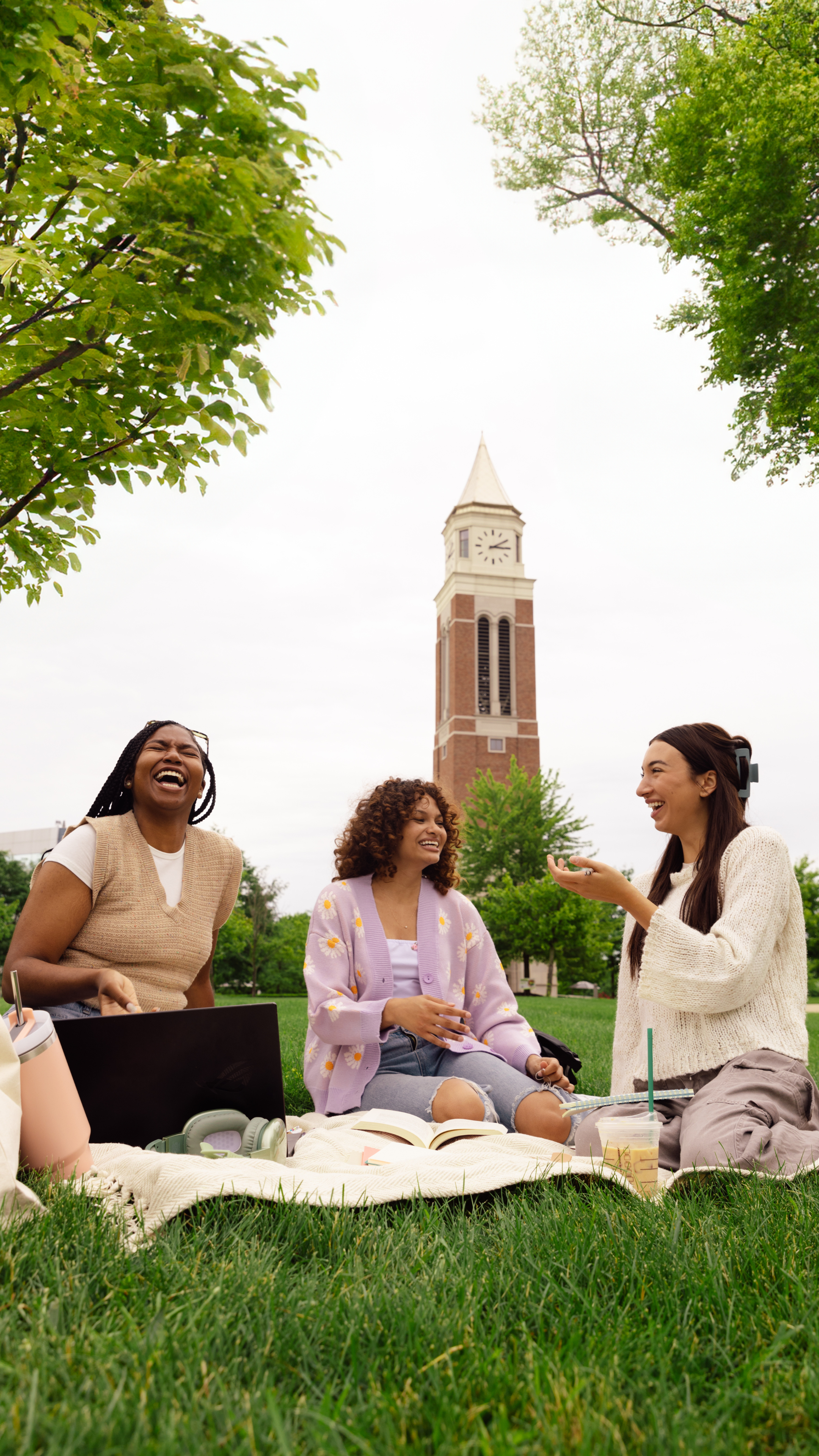 Three women sitting on a blanket in a park, laughing and talking. There are trees and a church steeple in the background.