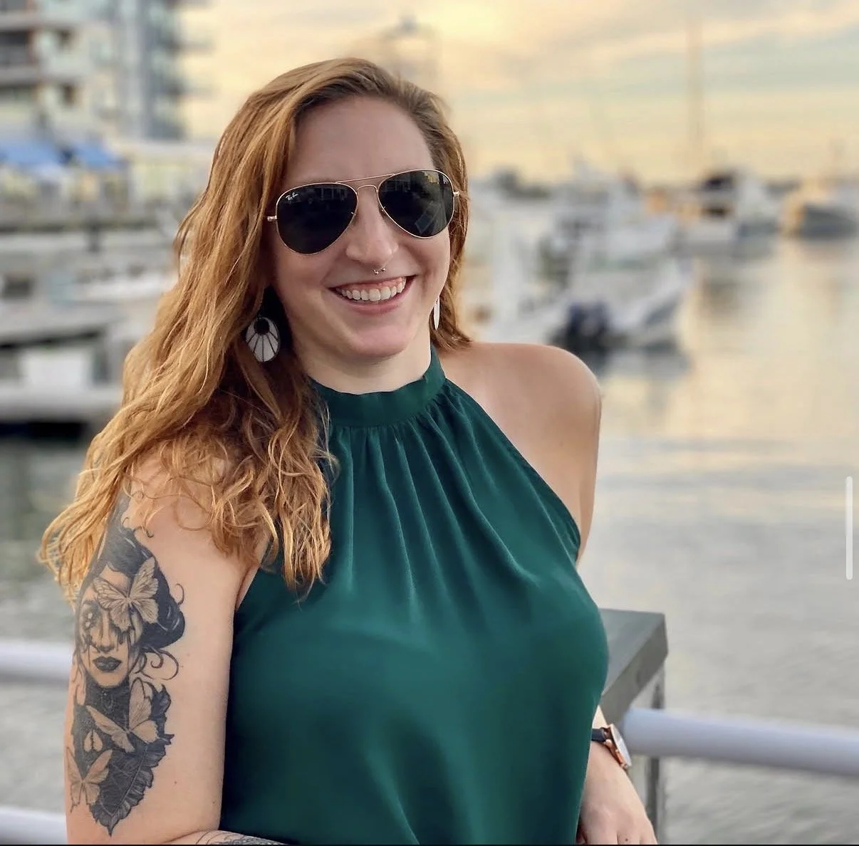 A woman with red hair, sunglasses, and a teal sleeveless top standing outdoors by a marina with boats at sunset.