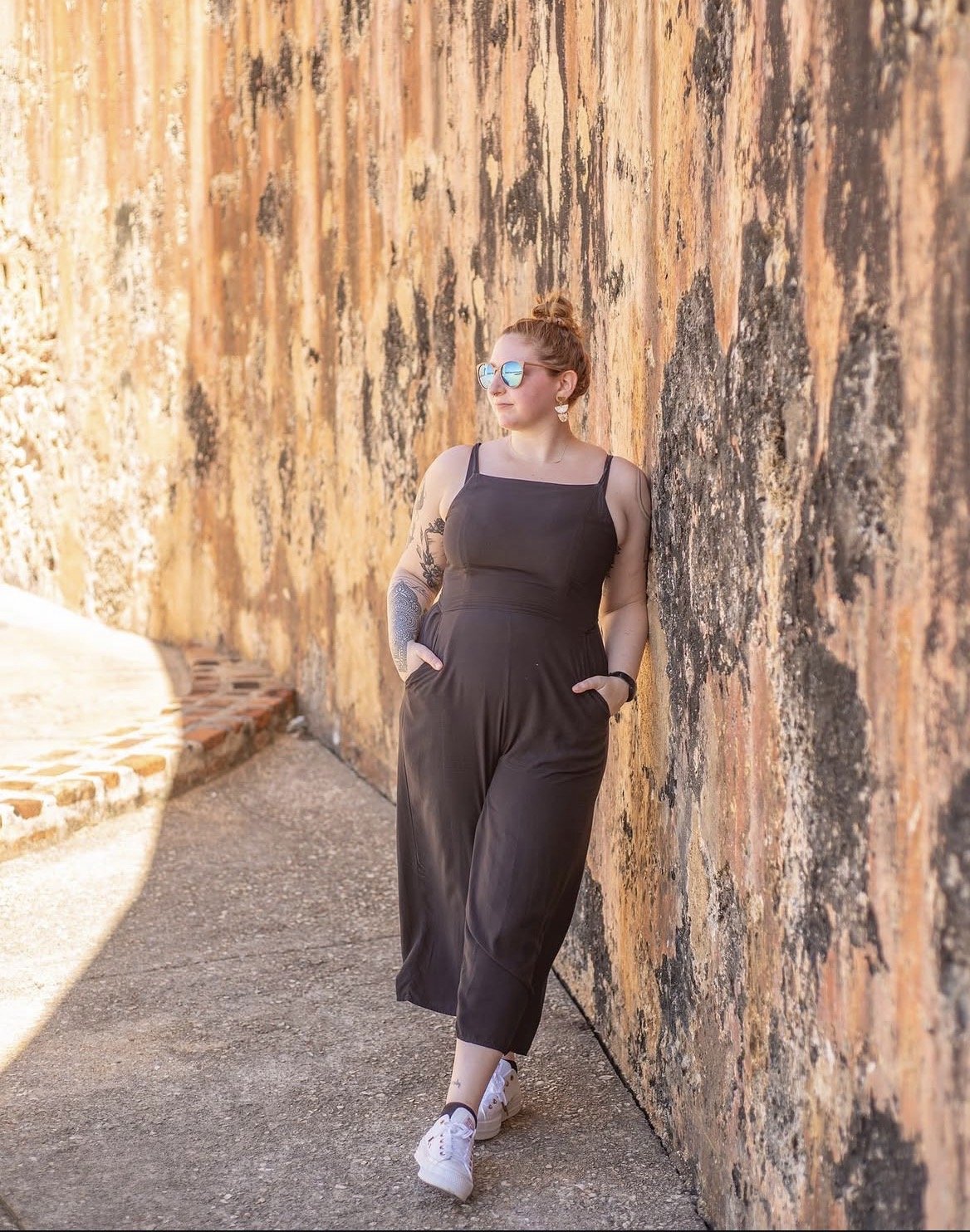 A woman with red hair styled in a bun, wearing reflective sunglasses and a black jumpsuit, stands against a textured stone wall with her hands in her pockets.