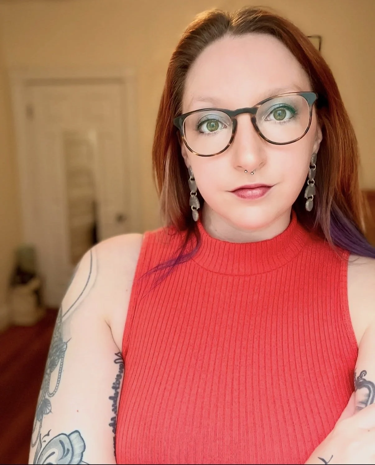 A woman with short red hair, glasses, and tattoos on her arm, wearing a red sleeveless top, taking a selfie in a warmly lit room.