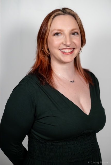 A woman with red hair, fair skin, and a nose piercing, smiling and wearing a black V-neck top against a plain light background.