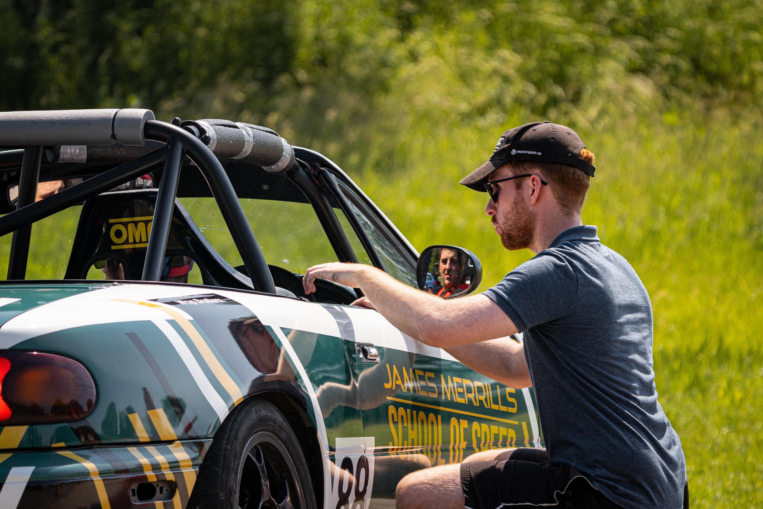 A young man in a gray t-shirt, black shorts, sunglasses, and a black cap kneels beside a race car, adjusting or inspecting it. The car has green, white, and yellow graphics with the text 'JAMES MERRILLS SCHOOL OF SPEED' on the side and the number 88. The background consists of green grassy trees.