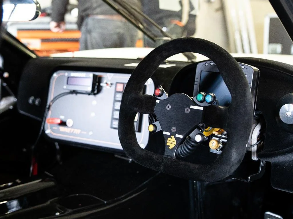 Inside a race car cockpit, featuring a black steering wheel with colored buttons and a digital display screen on the dashboard.