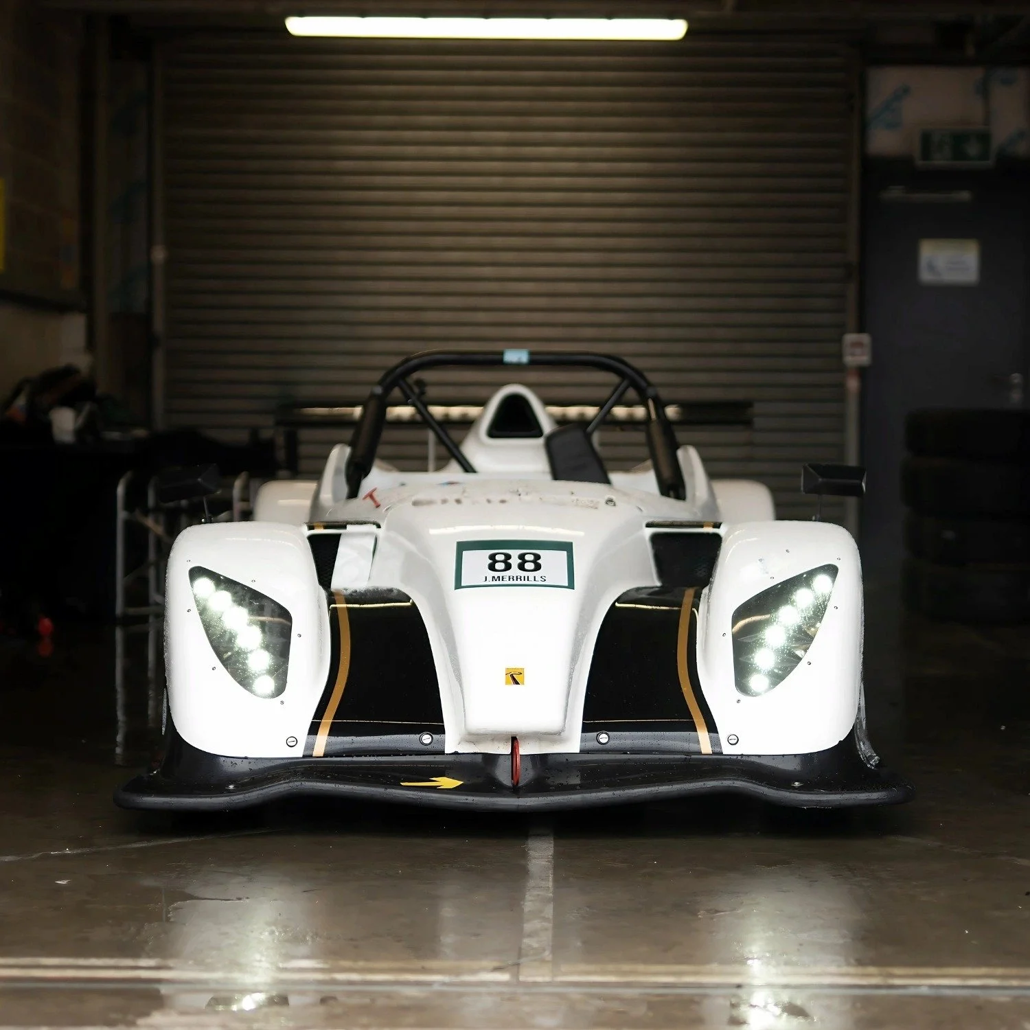 Front view of a white race car with LED headlights, black and gold accents, parked indoors in front of a metal garage door.