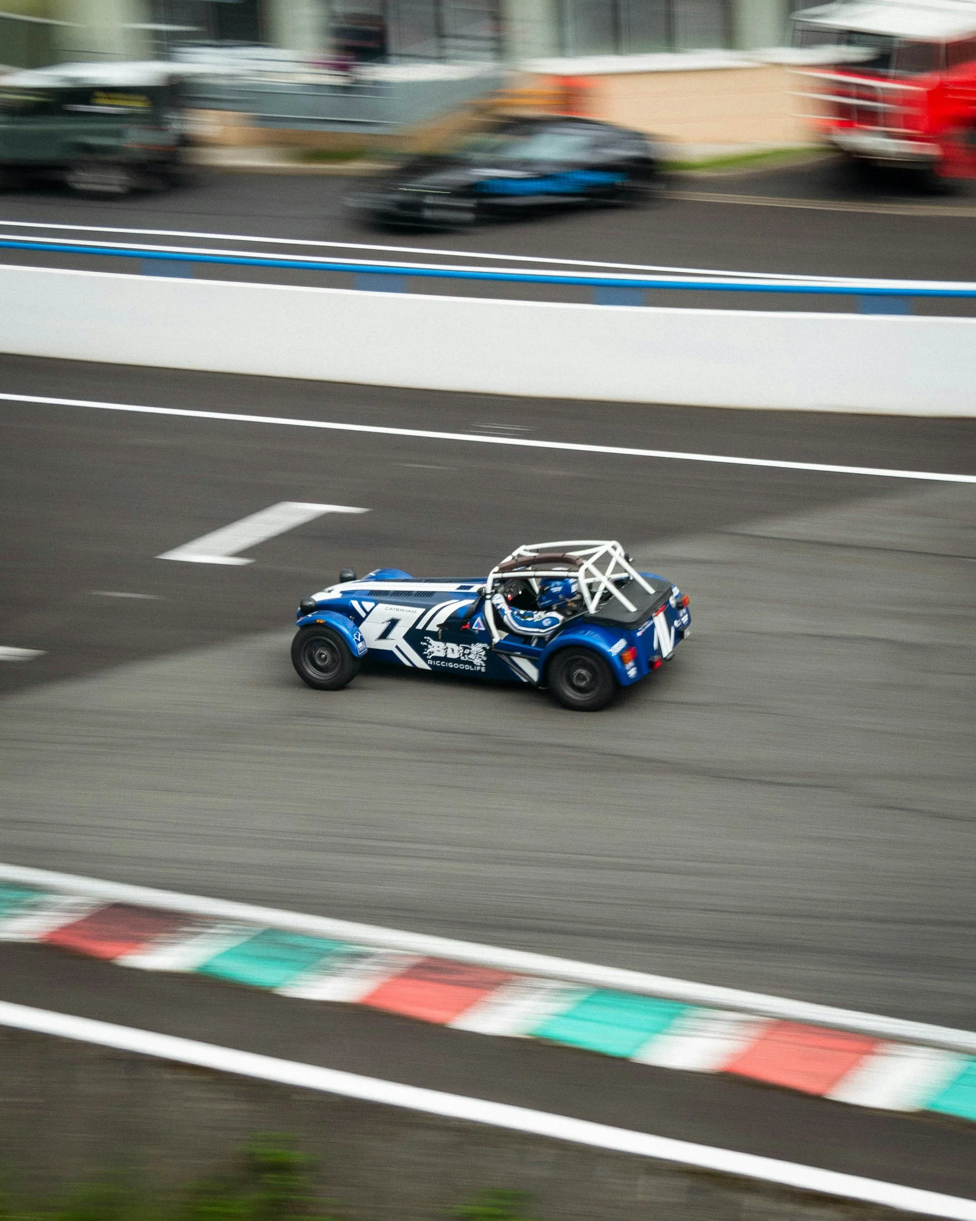 A blue race car speeding on a racetrack with blurred background showing other vehicles and barriers.