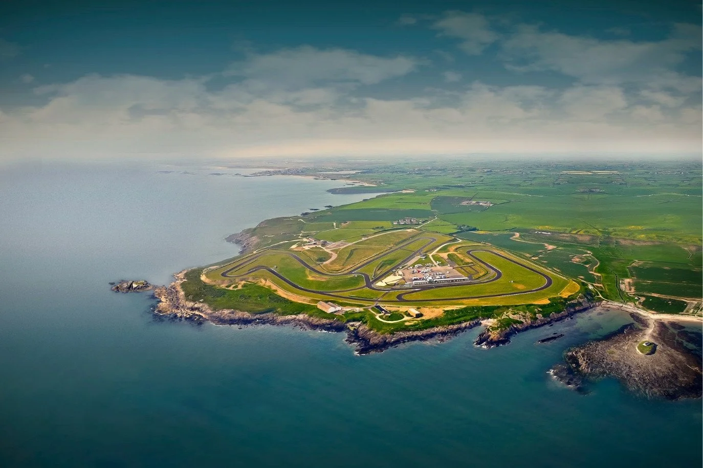 Aerial view of Anglesey race track on a green peninsula with winding roads, surrounded by ocean and farmland.