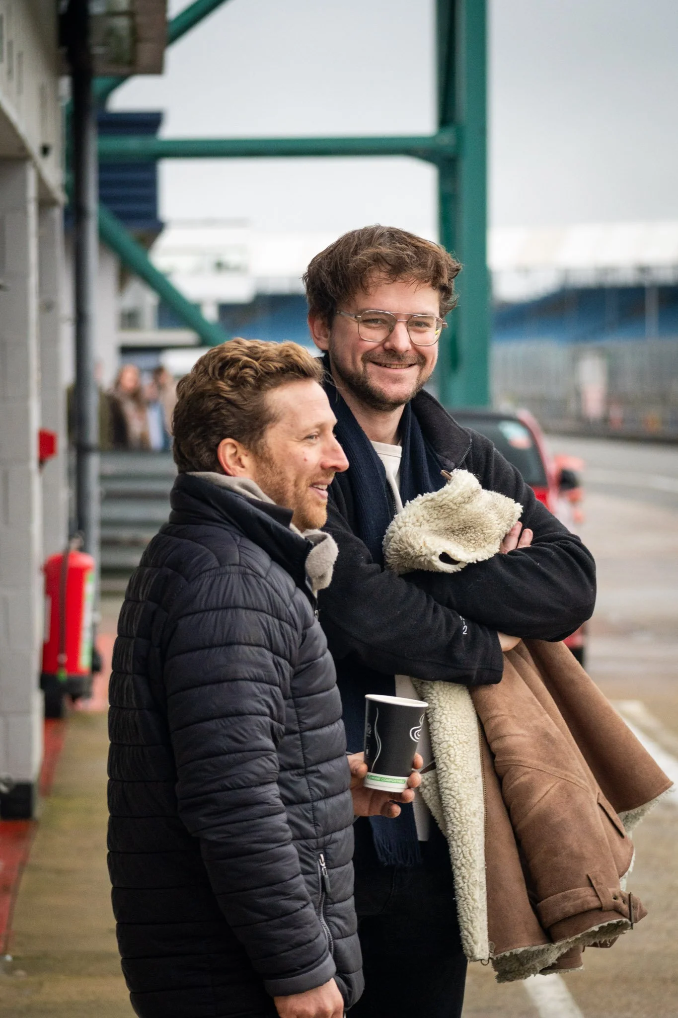 Two men standing in Silverstone Pit Lane, smiling and chatting. One man is holding a coffee cup, and the other is holding a coat.