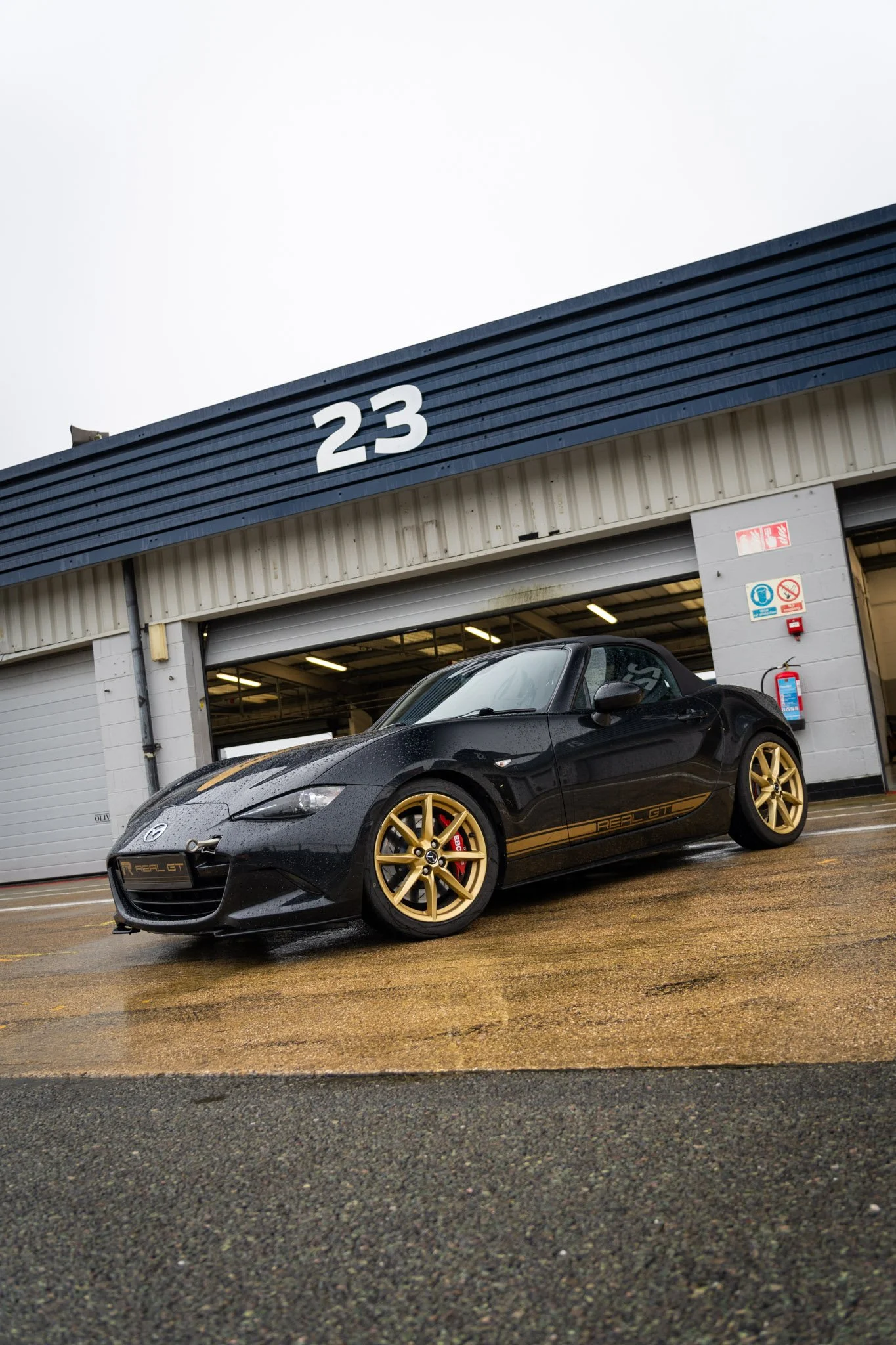 A black and gold Mazda MX5 parked outside garage number 23 in Silverstone's National Pit Lane, on a rainy day during a driving experience