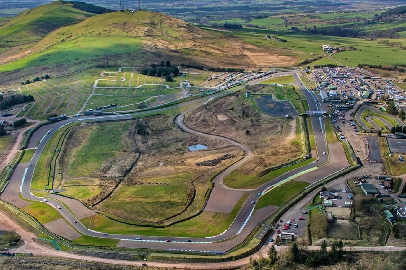 An aerial view of Knockhill race track, Dunfermline, Scotland, with winding asphalt track, surrounded by grassy areas, parking lots filled with cars, and hills in the background.