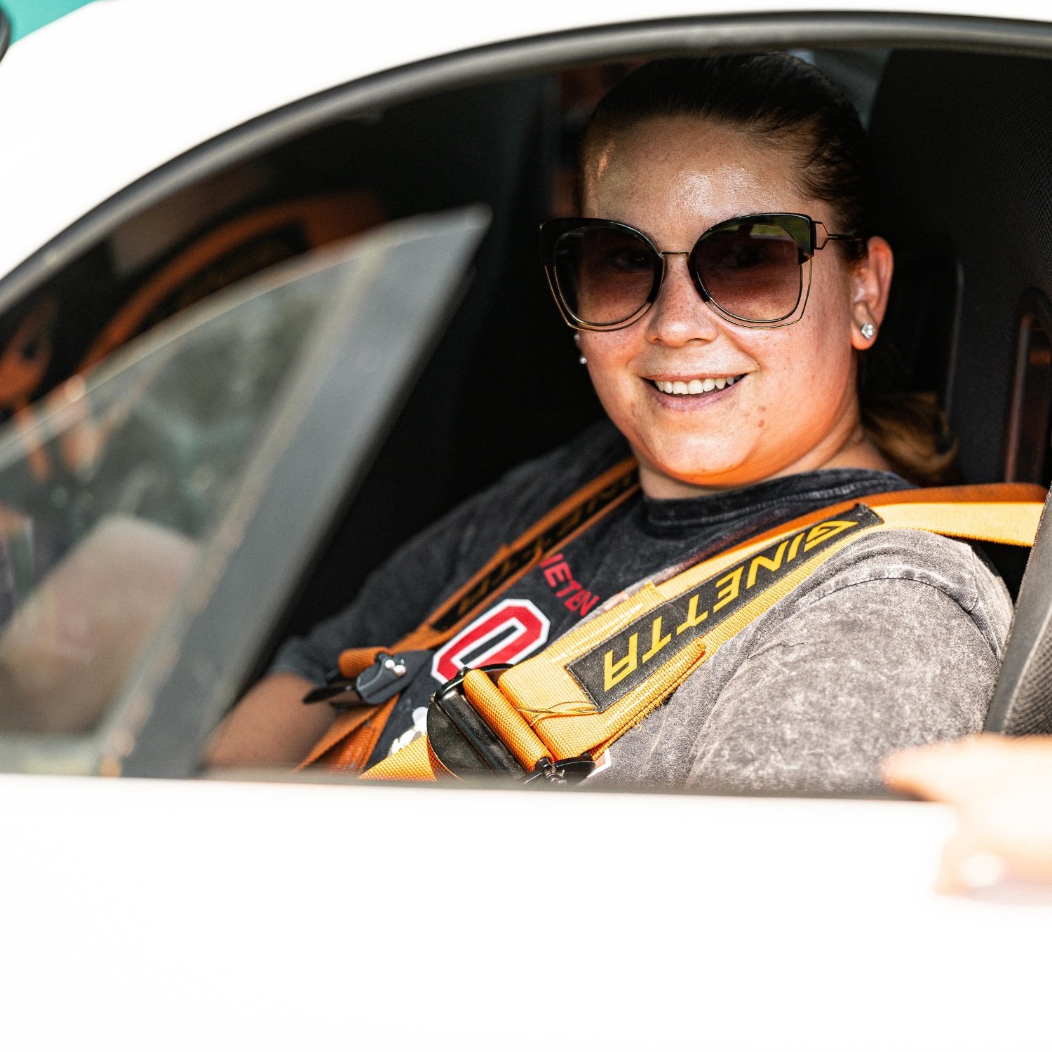 Smiling woman in sunglasses wearing a racing harness sitting in a race car.