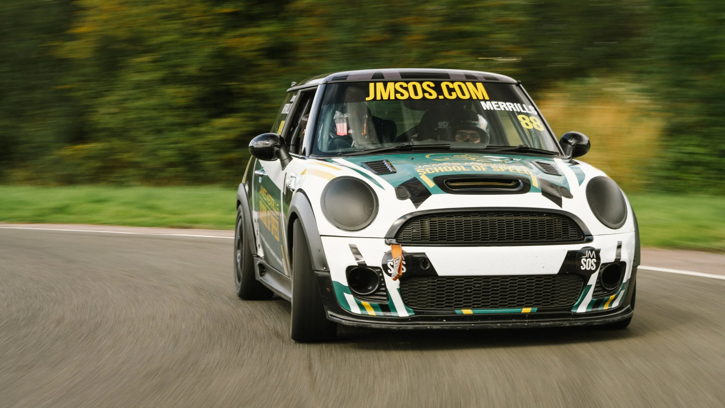 A racing Mini Cooper driving fast on a curved road with a blurred forest background.
