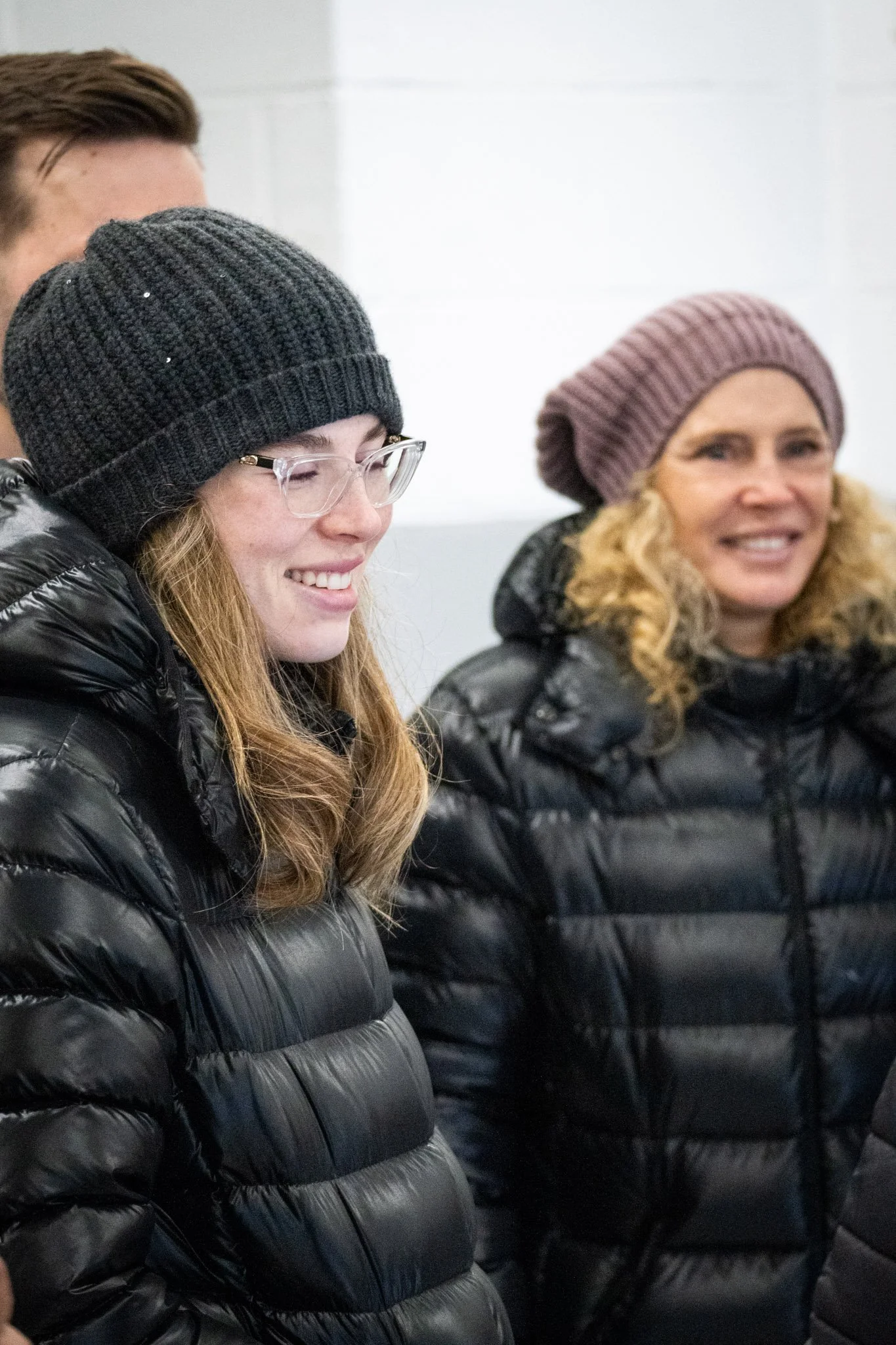 Smiling woman in glasses and black beanie, wearing a black puffer jacket, standing indoors next to another woman with curly blonde hair in a pink beanie, also wearing a black puffer jacket.