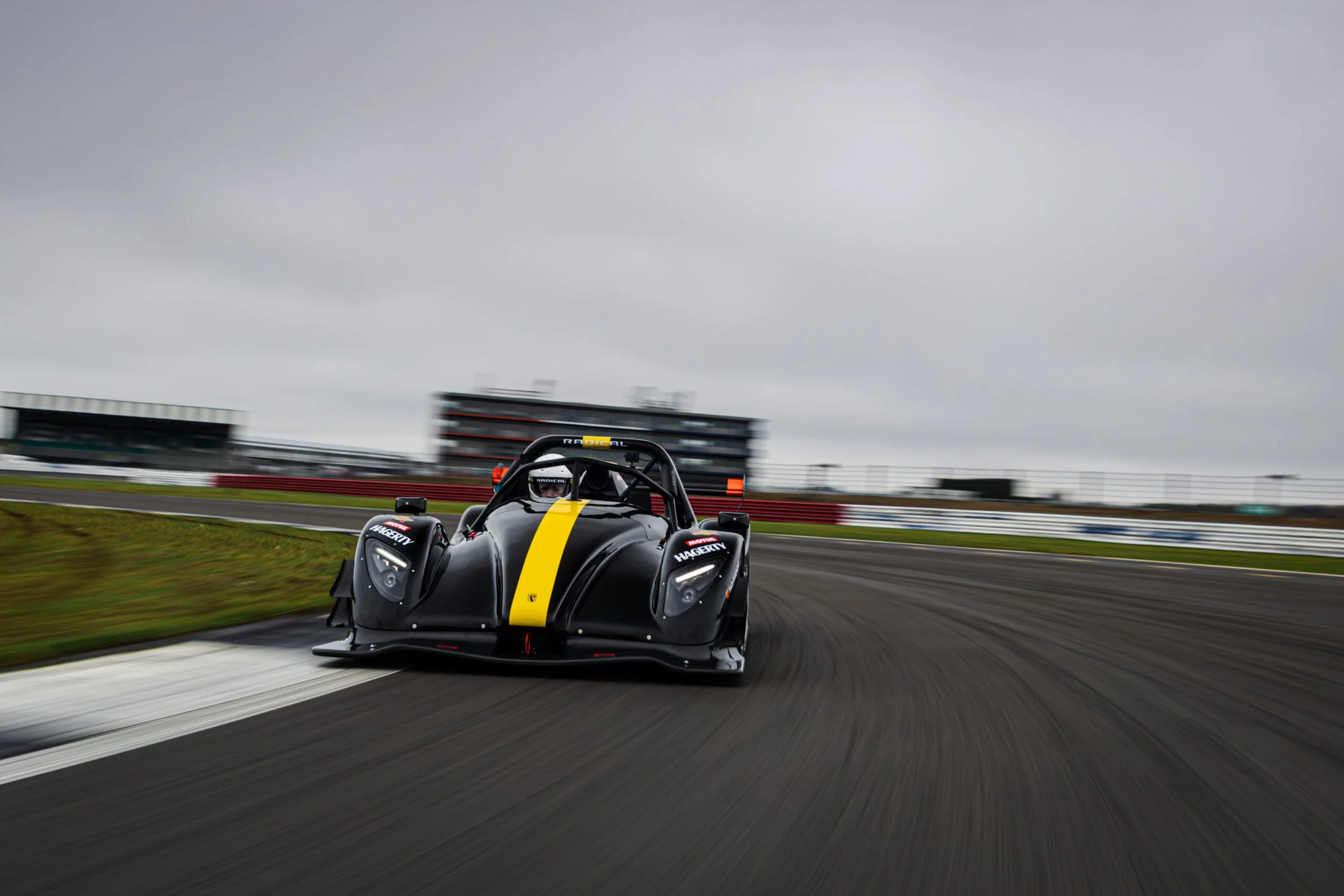 A black race car with yellow stripe on a race track during overcast weather, curving along the track with a blurred grandstand and cloudy sky in the background.