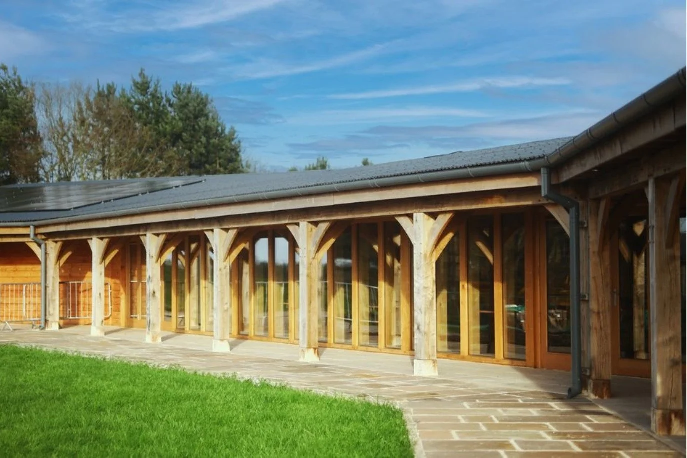 A building with a wooden structure and glass doors, a sloped roof with solar panels, a stone pathway, and a green lawn, with trees and a blue sky in the background. This is the hospitality suite at Castle Combe race track, near Bristol.