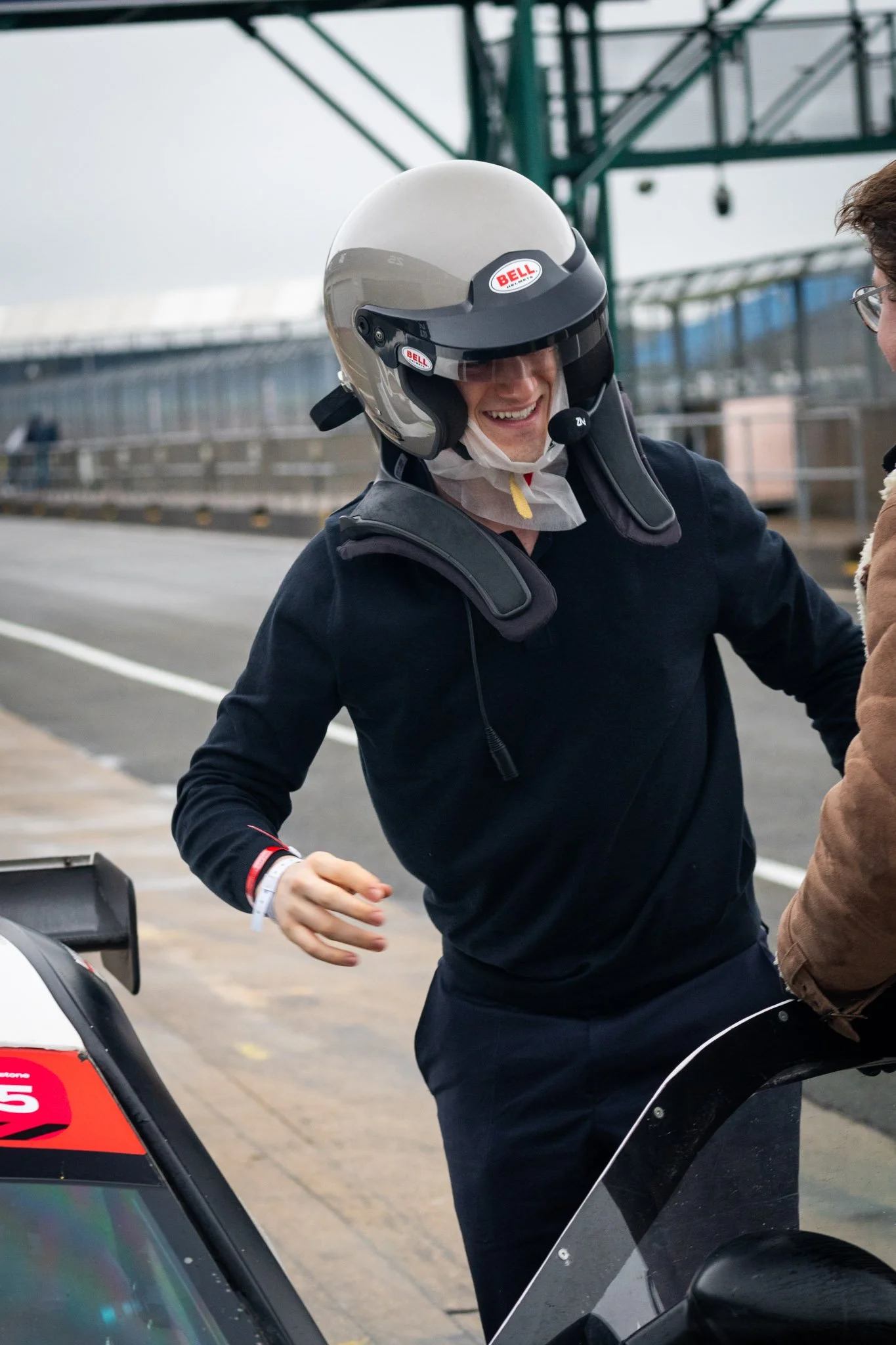 A person wearing a racing helmet, smiling, standing next to a racing race car, in the pit lane at Silverstone