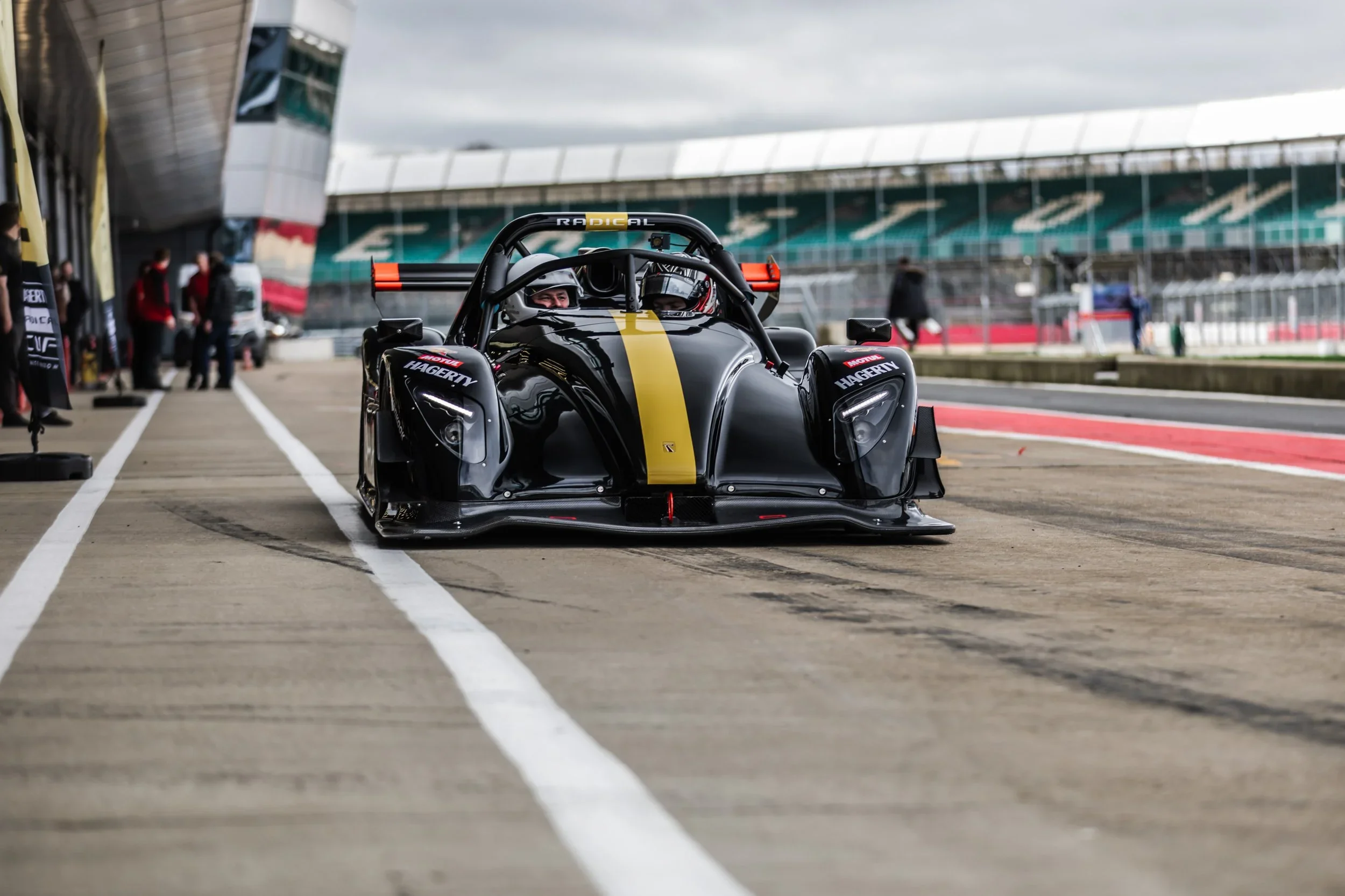 A sleek black race car with gold accents and the word 'RADICAL' on the windshield, parked in a pit lane at a race track with some people and structures in the background.