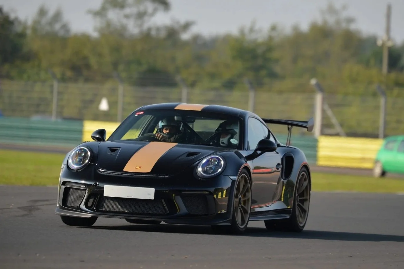 A black sports car with tan racing stripes on a race track, with a driver wearing a helmet inside. The car has a rear wing and is in motion, at Castle Combe race track, near Bristol.
