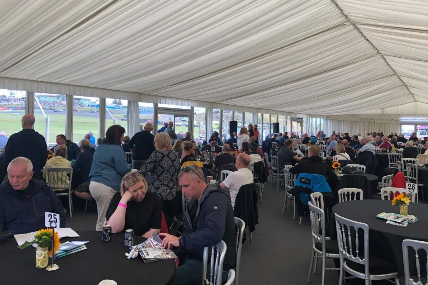 People sitting and standing inside a large tent, attending an event near a race track. Round tables with sunflower centerpieces, some with magazines and drinks.