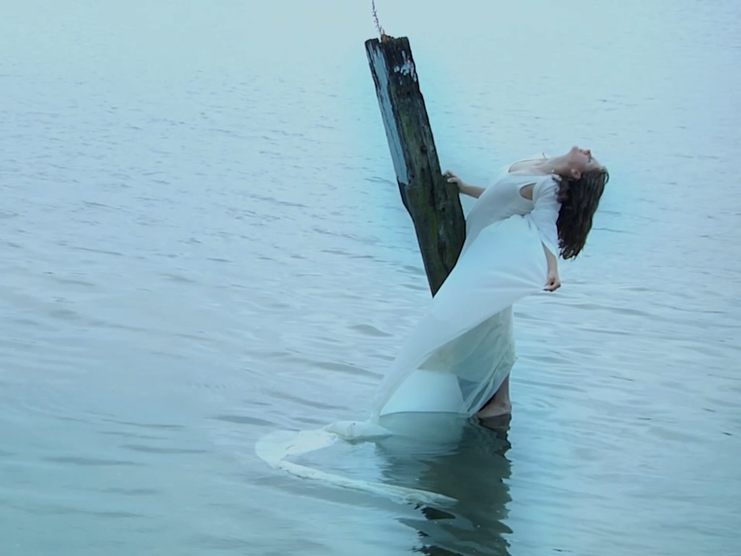 A woman in a white dress is standing in water, leaning back with her head tilted upwards, holding onto a wooden post.