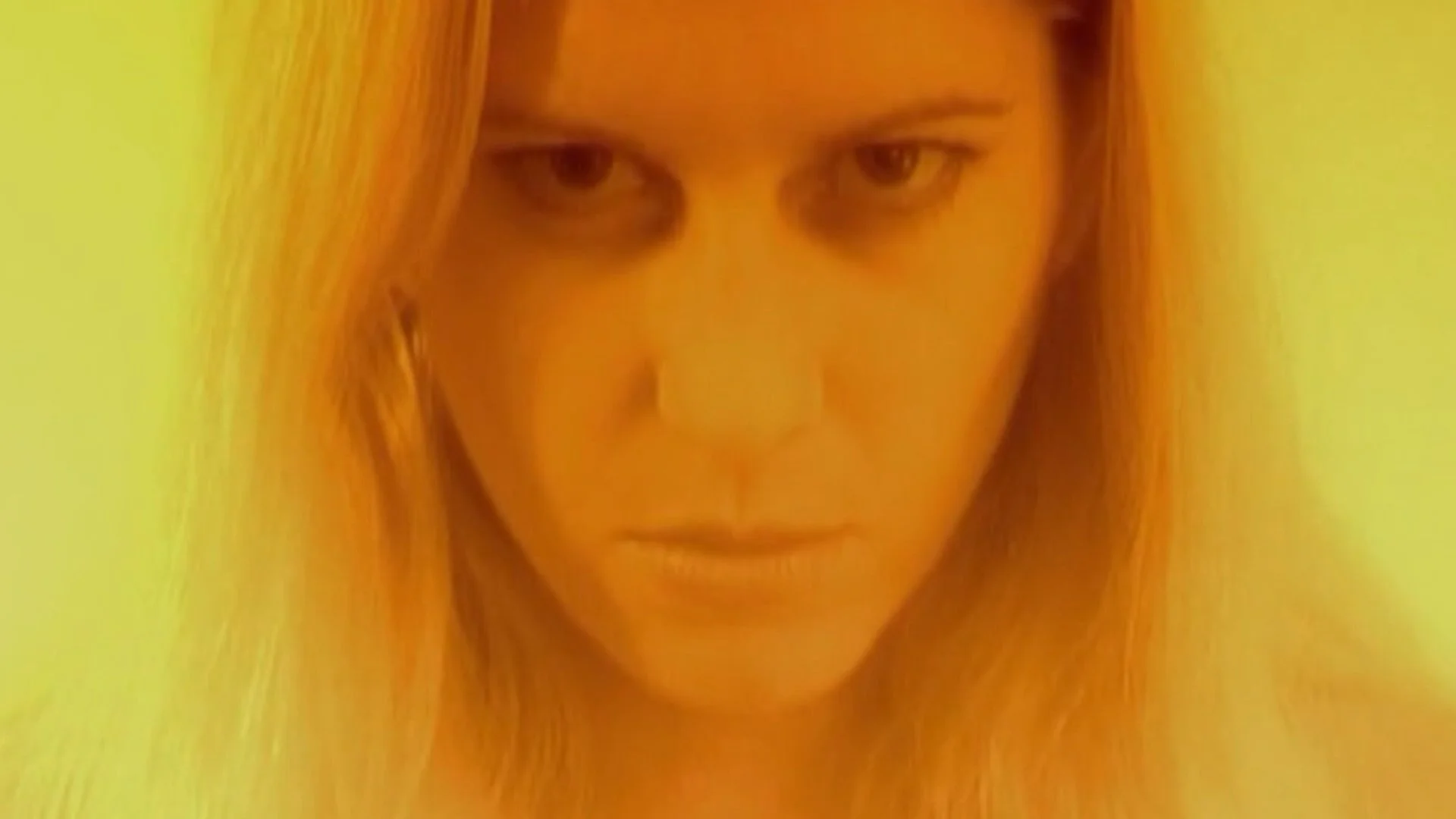 Close-up of a woman with long blonde hair and intense expression, looking directly at the camera against a plain background.