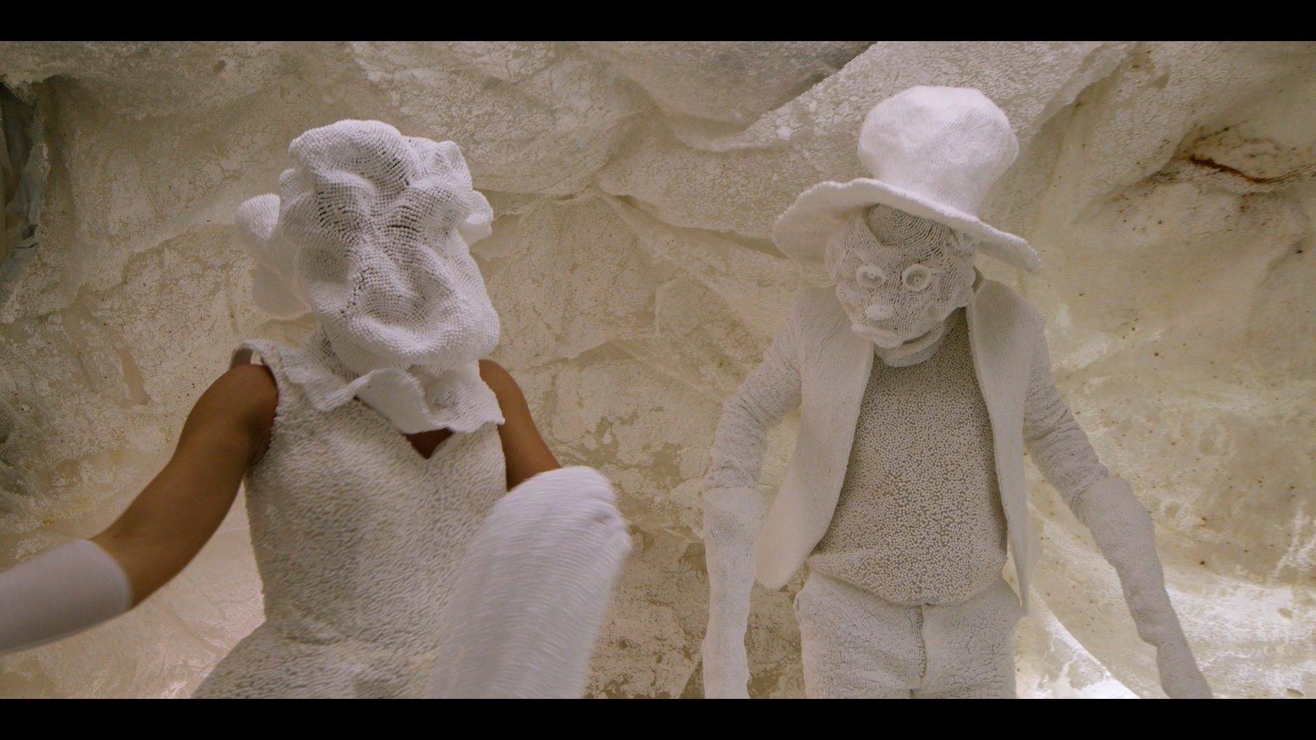 Two sculptures of people made from white textured material, one with a hat, in a cave-like space.