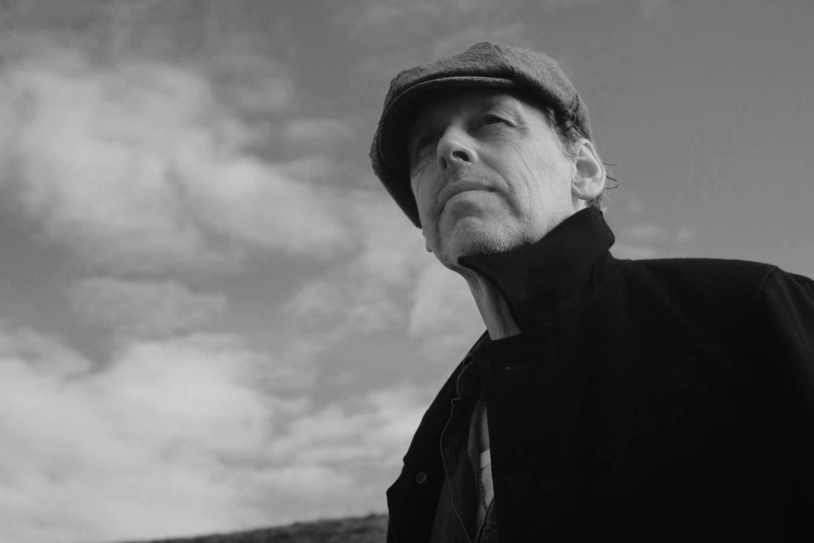 Black and white photo of an older man wearing a flat cap and a jacket, looking into the distance against a cloudy sky.