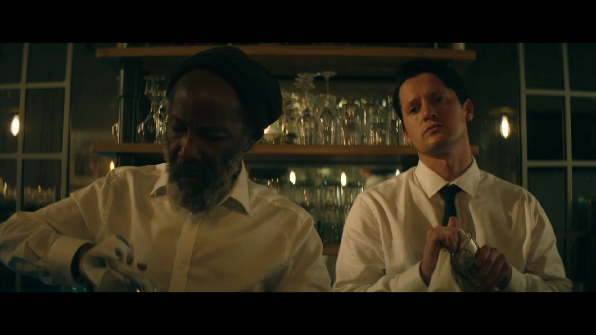 Two men in white shirts and black ties behind a bar, one with a dark complexion and beard, the other with a light complexion and dark hair. The man on the left is looking down, and the man on the right is holding a cloth and looking pensively.