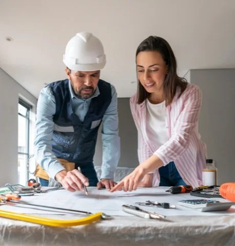 A male construction worker wearing a white hard hat and blue vest reviewing blueprints with a smiling woman in a pink and white striped shirt at a table.