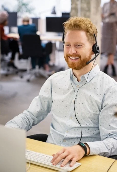 A man with red hair and a beard wearing a light-colored, dotted shirt, headset, and smartwatch, smiling while working on a computer in an office setting.