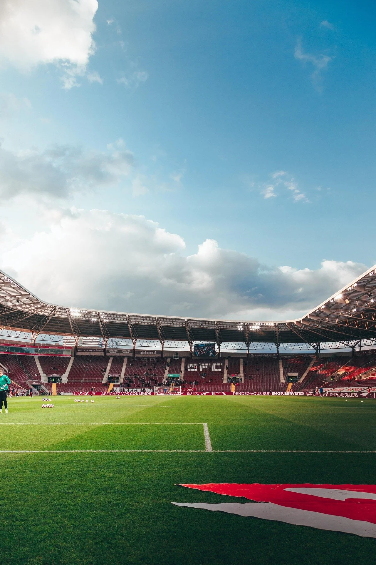 Empty soccer stadium with bright green field under a blue sky with clouds, banners and advertising around the perimeter, and a few players warming up.