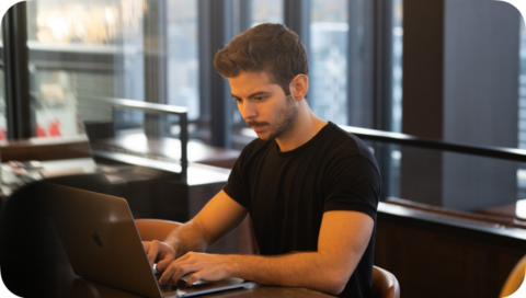 A young man working on a laptop at a table in a cafe or restaurant with large windows.