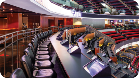 Empty row of black chairs and desks inside a stadium or arena with red and yellow seating areas in the background.