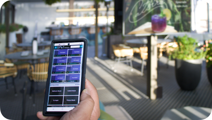 Person holding a smartphone with SAVI control interface open on an outdoor patio, with tables, chairs, and a sign that has a purple drink on it in the background.