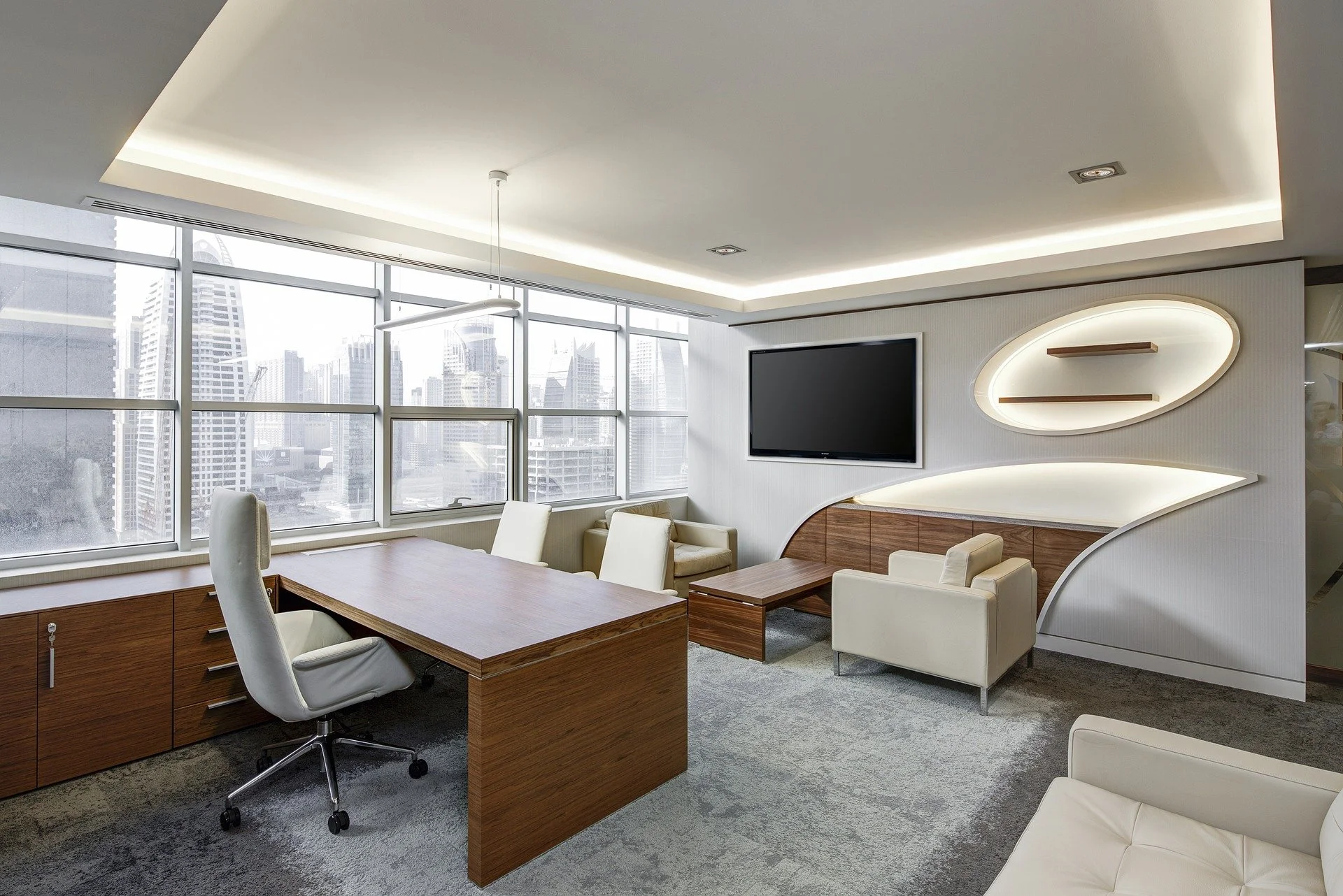 Modern office with a large window view of a city skyline, a wooden desk with a white office chair, a TV on the wall, and contemporary seating with beige and white furniture.