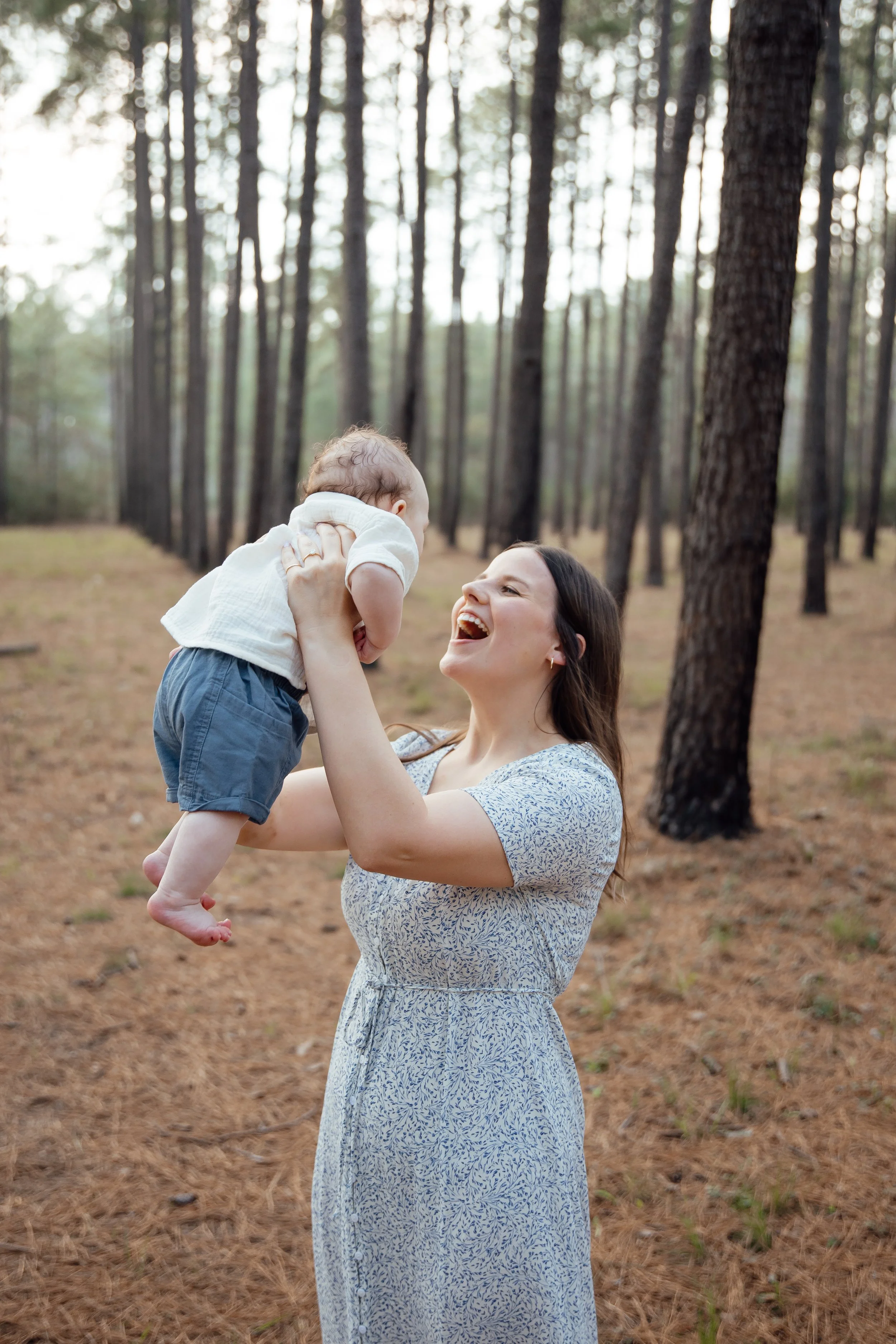 A woman joyfully lifting a baby in a forest with tall trees and pine needles on the ground.