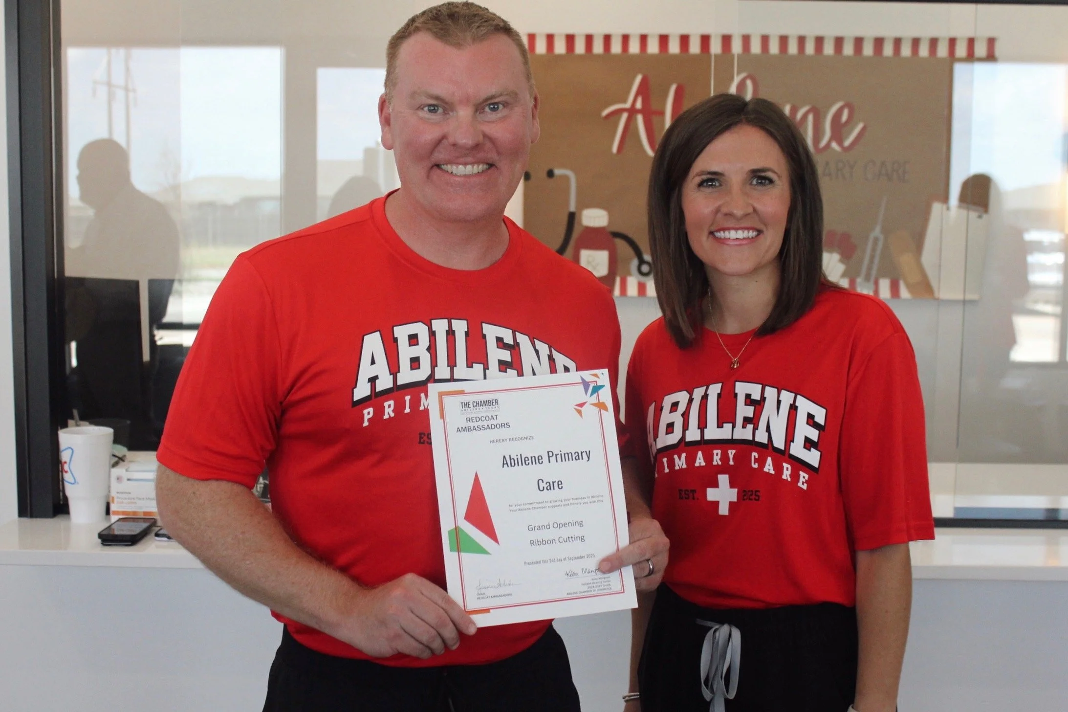 Chris Shanklin and Caitlin Blair wearing Abilene Primary Care shirts holding a certificate during a ribbon-cutting event at the clinic.