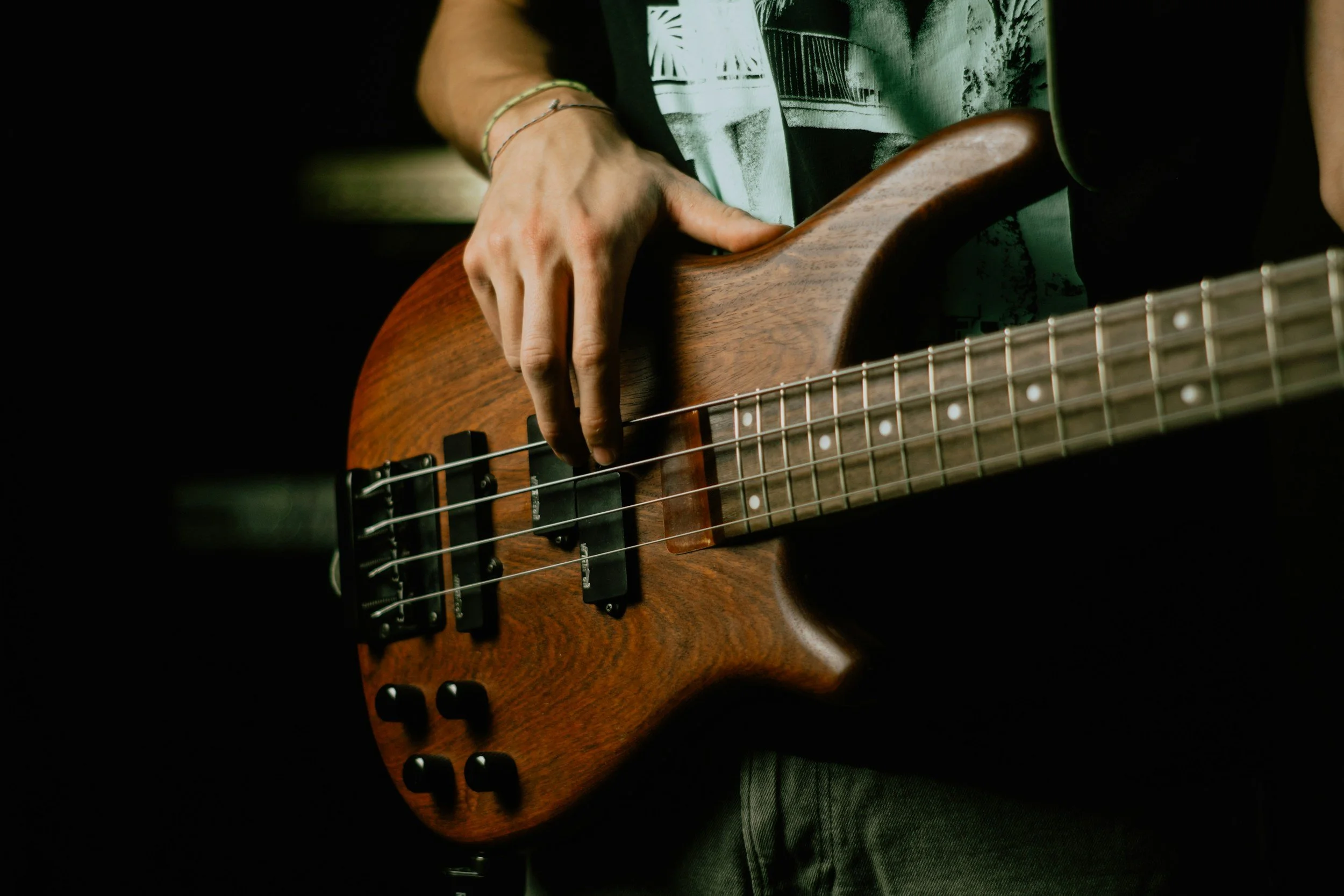 Close-up of a person playing a wooden electric bass guitar with four strings, black pickups, and control knobs, against a dark background.
