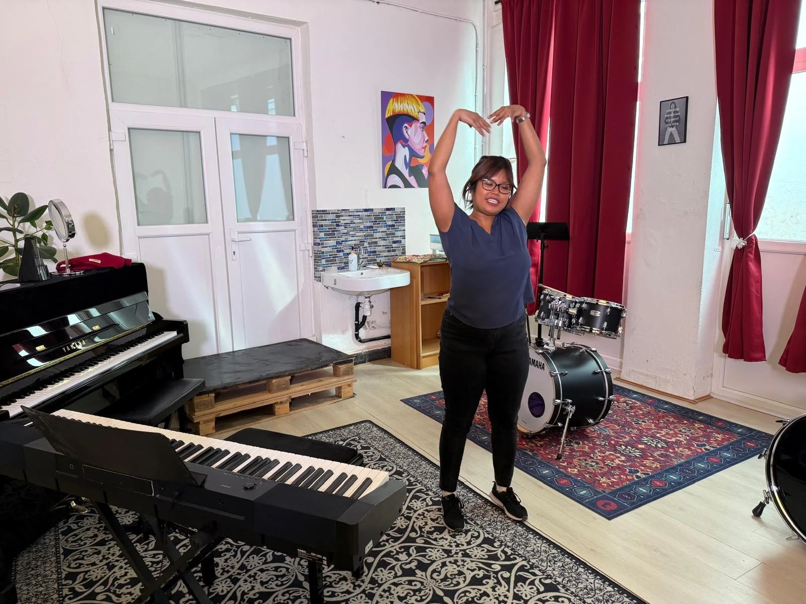 A woman with glasses and dark hair in a music studio, standing on a rug, dancing with her arms raised above her head, surrounded by musical instruments including a black piano, a drum set, and a black keyboard.
