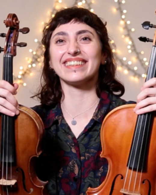 A woman smiling and holding two violins in front of a background with string lights.