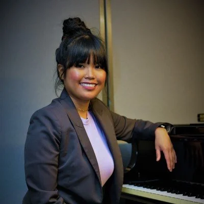 Smiling woman with dark hair styled in a bun, wearing a gray blazer, sitting beside a piano in an indoor setting.