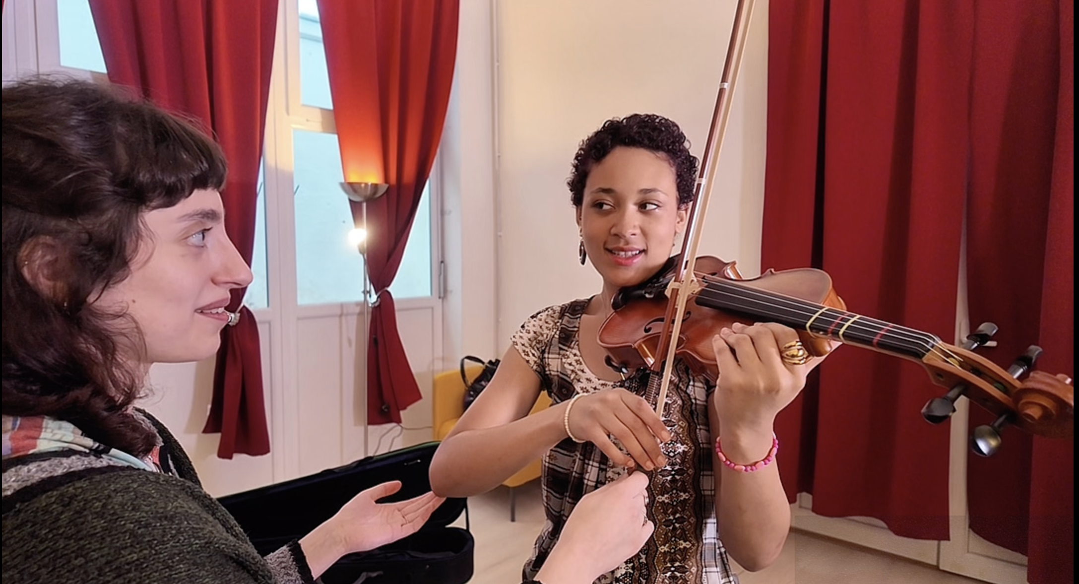 A young woman playing a violin while another woman assists her during a music lesson in a room with red curtains and natural light.