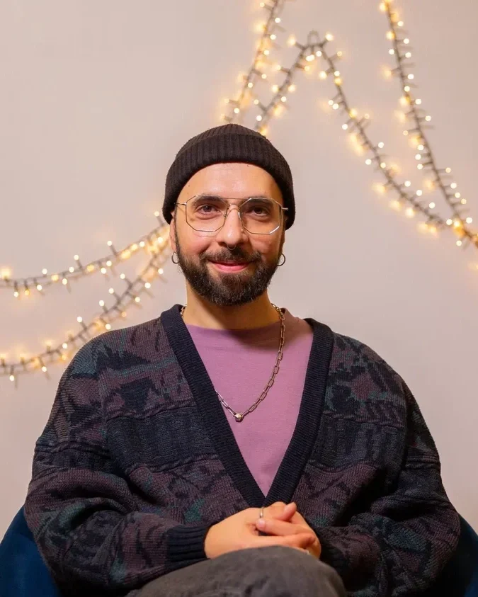 A man with glasses and a beard wearing a black beanie, purple shirt, and patterned cardigan, sitting in front of a wall decorated with string lights.