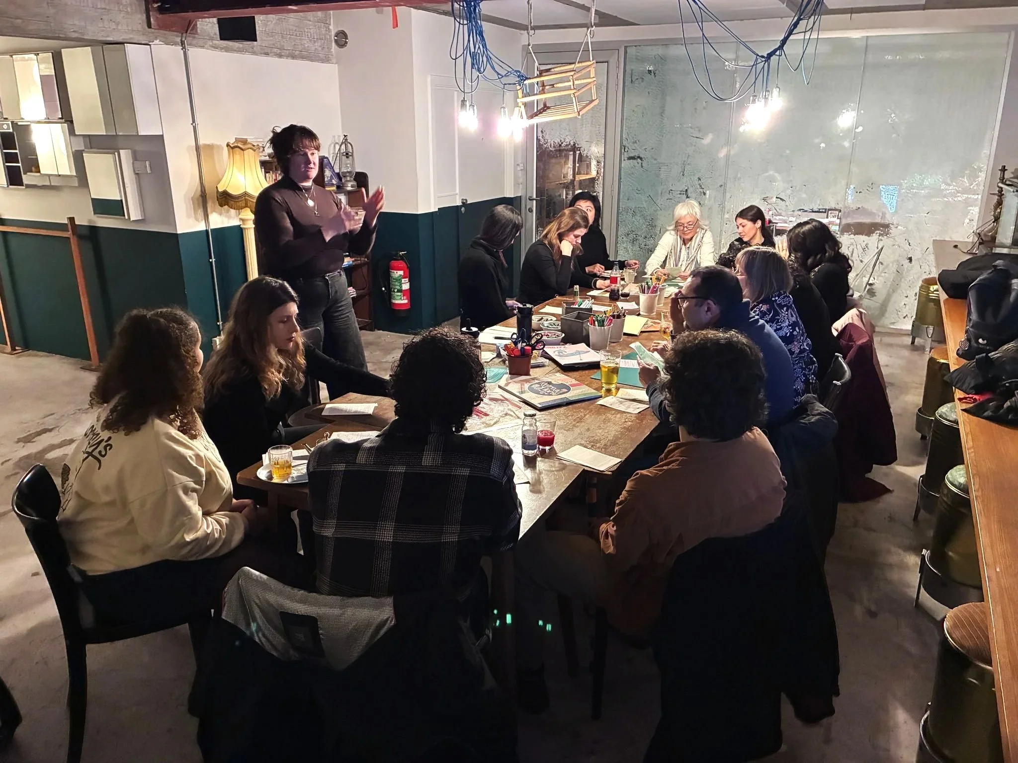 A woman is standing and speaking to a group of people in a dimly lit room with industrial decor, wooden tables, and colorful books and supplies on the tables. Some attendees are listening, with one person looking down at their phone. The room has exposed pipes, a large glass window, and a mix of vintage and modern furnishings.