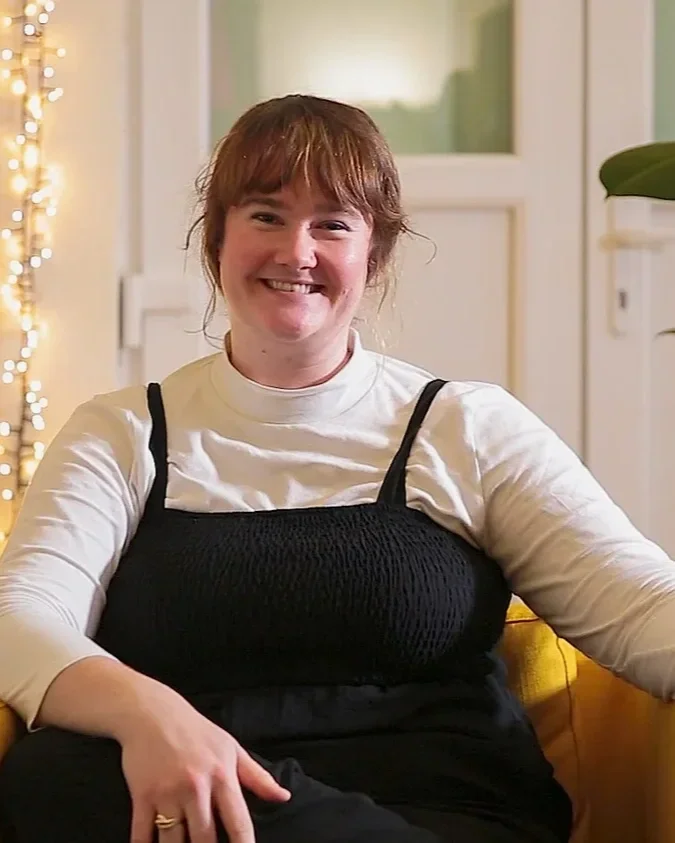 A smiling woman with short auburn hair, wearing a white long-sleeve top underneath a black sleeveless top, sitting on a yellow couch in a cozy, warmly lit room.