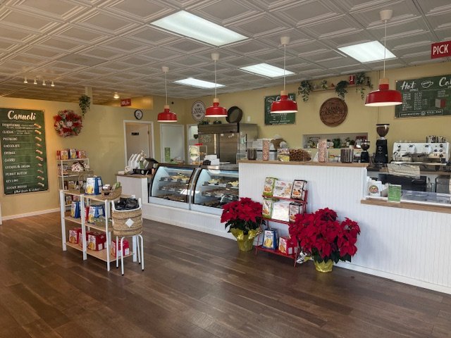 Coffee shop with a display case for baked goods, red pendant lights, Christmas decorations, and a menu board, with a small table of snacks and magazines in the foreground.