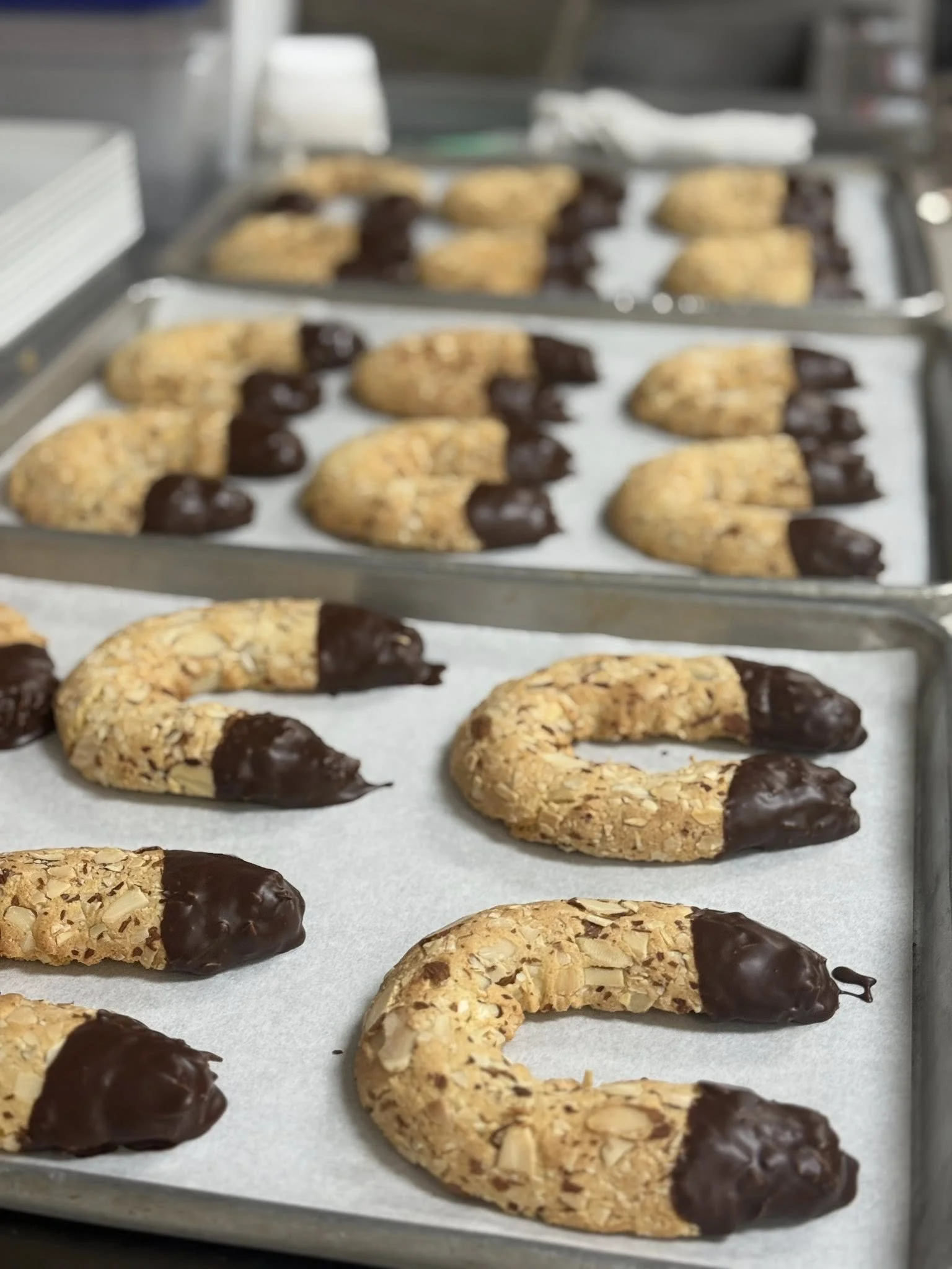 Chocolate-dipped almond cookies shaped like crescent moons on baking sheets lined with parchment paper.